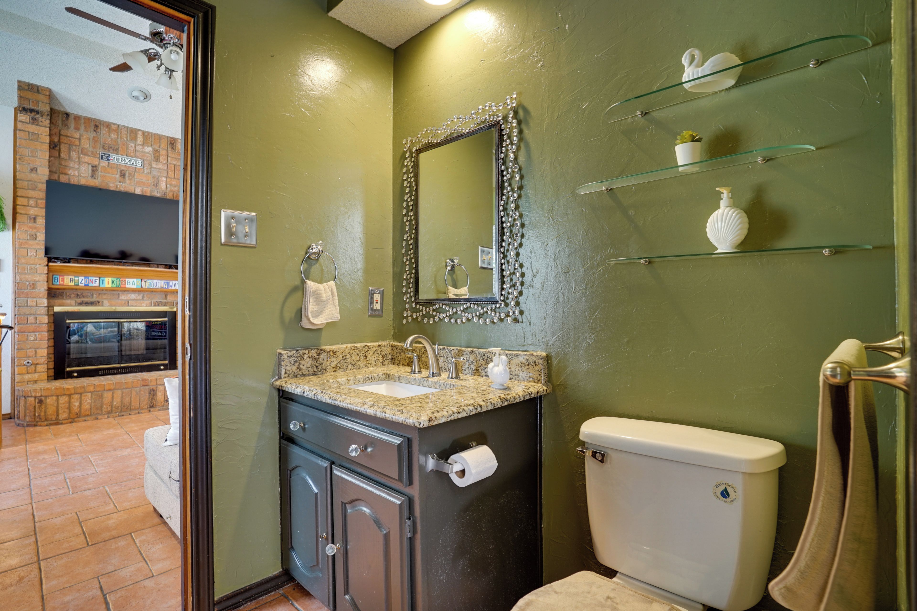 A bathroom with a granite countertop sink, a toilet, and a glass shelf with toiletries.
