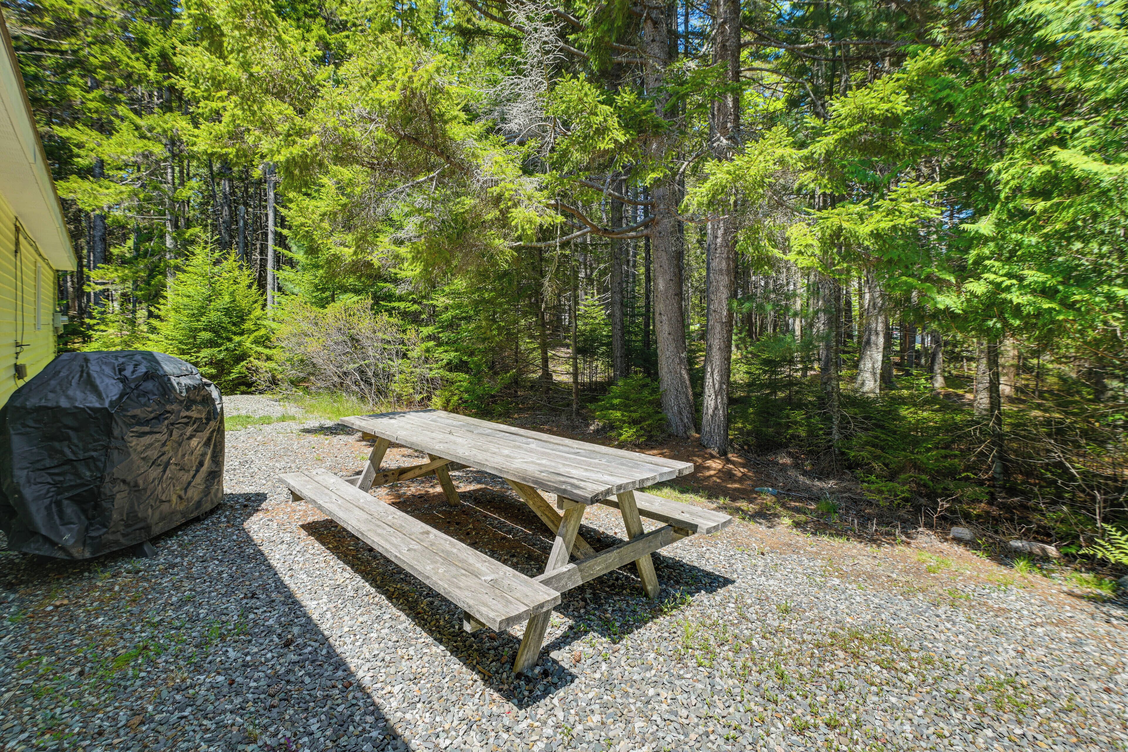 Ein Holztisch für ein Picknick in bewaldeter Landschaft, mit einem überdachten Grill und einem gelben Gebäude im Hintergrund.