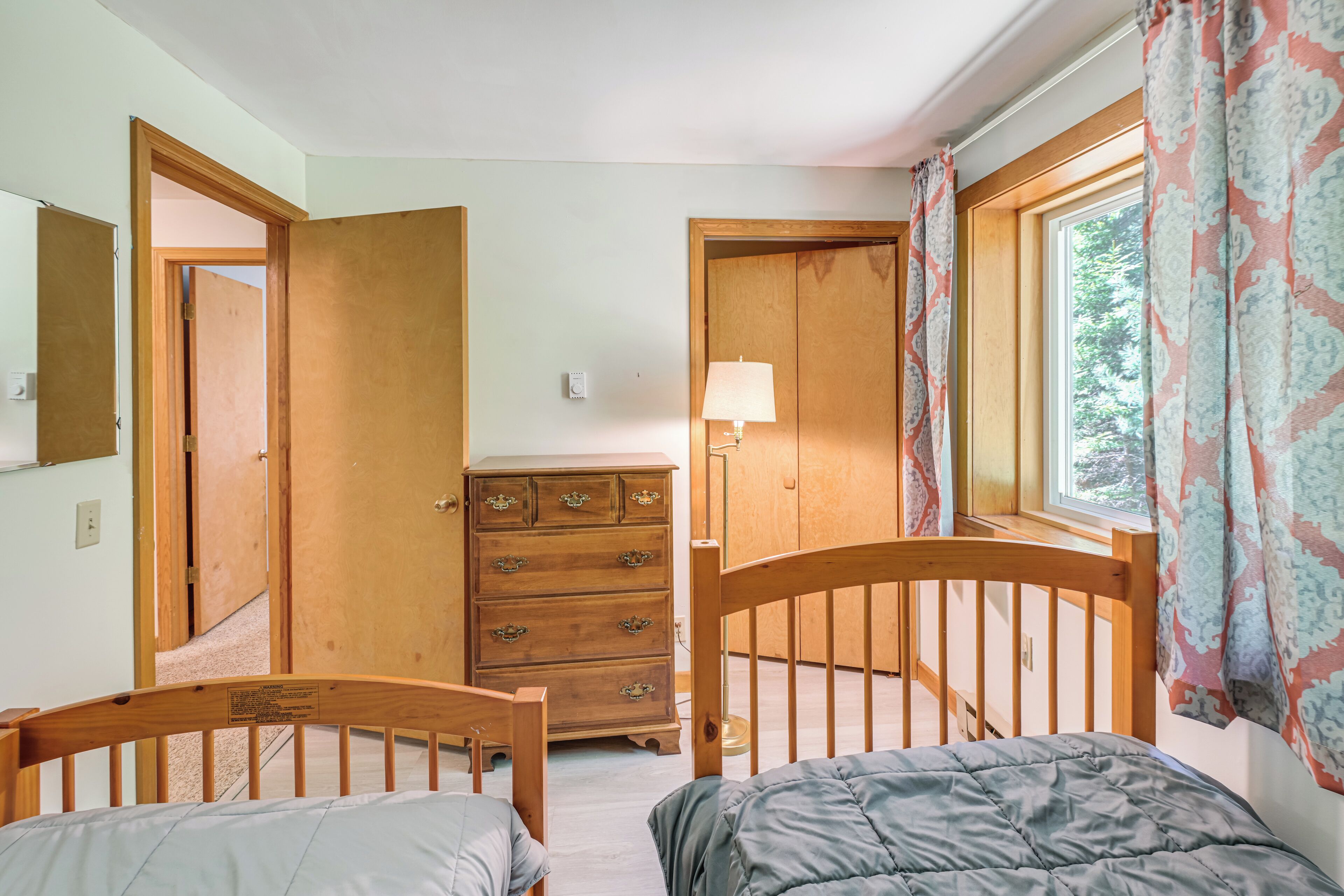 Un dormitorio con dos camas de madera, un aparador, una ventana con cortinas y una puerta que da a otra habitación.