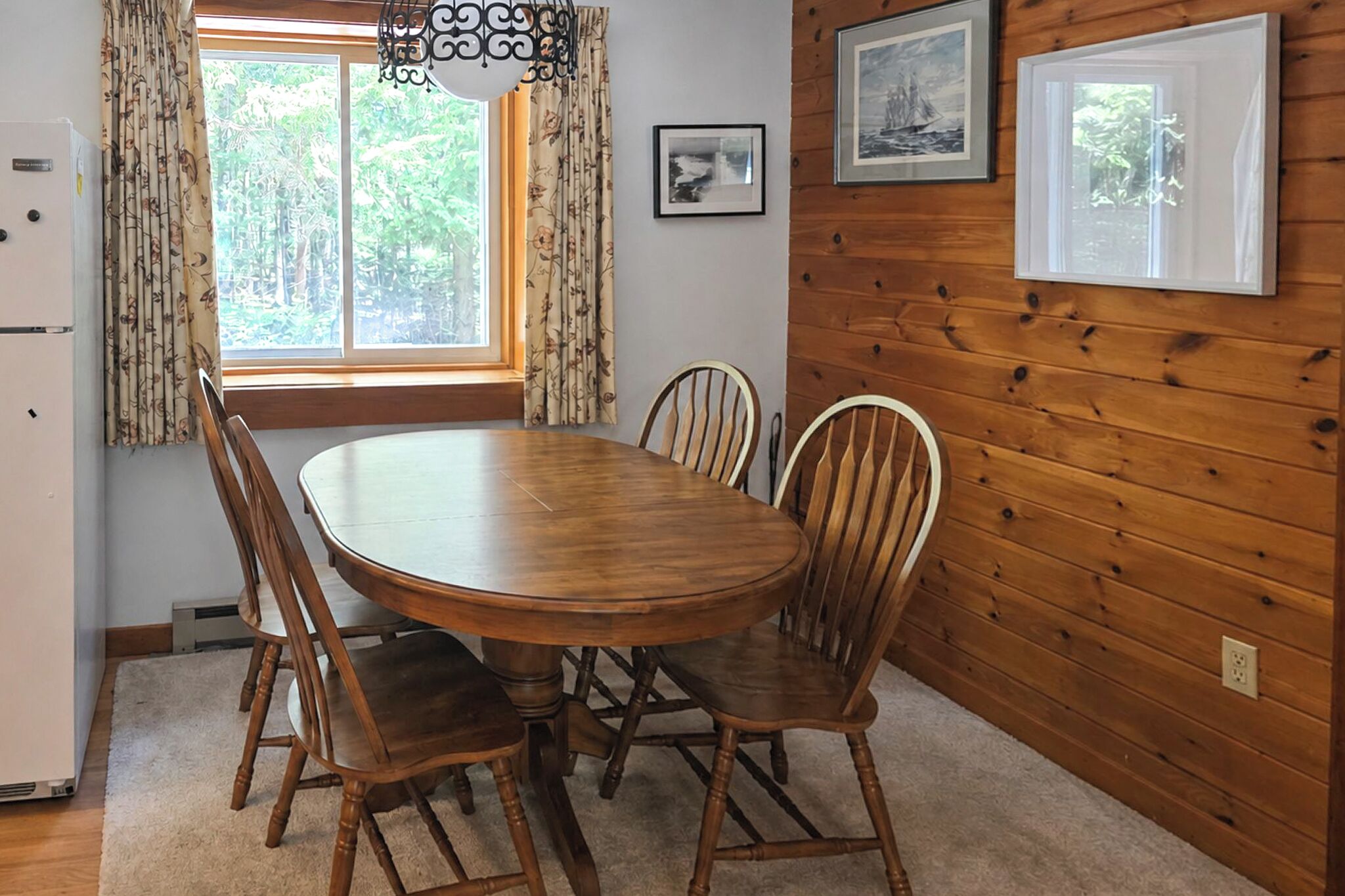 A wooden-paneled room with a round dining table, chairs, a refrigerator, and a window with curtains.