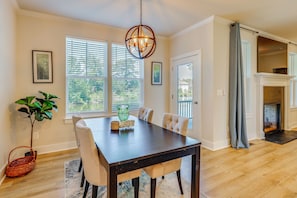 Uma sala de jantar com mesa de madeira escura, cadeiras estofadas brancas, luminária pendente e vista para o exterior através de grandes janelas.
