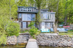 A two-story cabin with a stone staircase leading to a wooden deck, surrounded by trees.