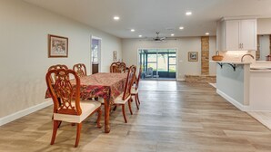 Un coin repas avec une table dressée pour six personnes, des chaises en bois, un ventilateur de plafond, un éclairage encastré et une vue sur l’extérieur.