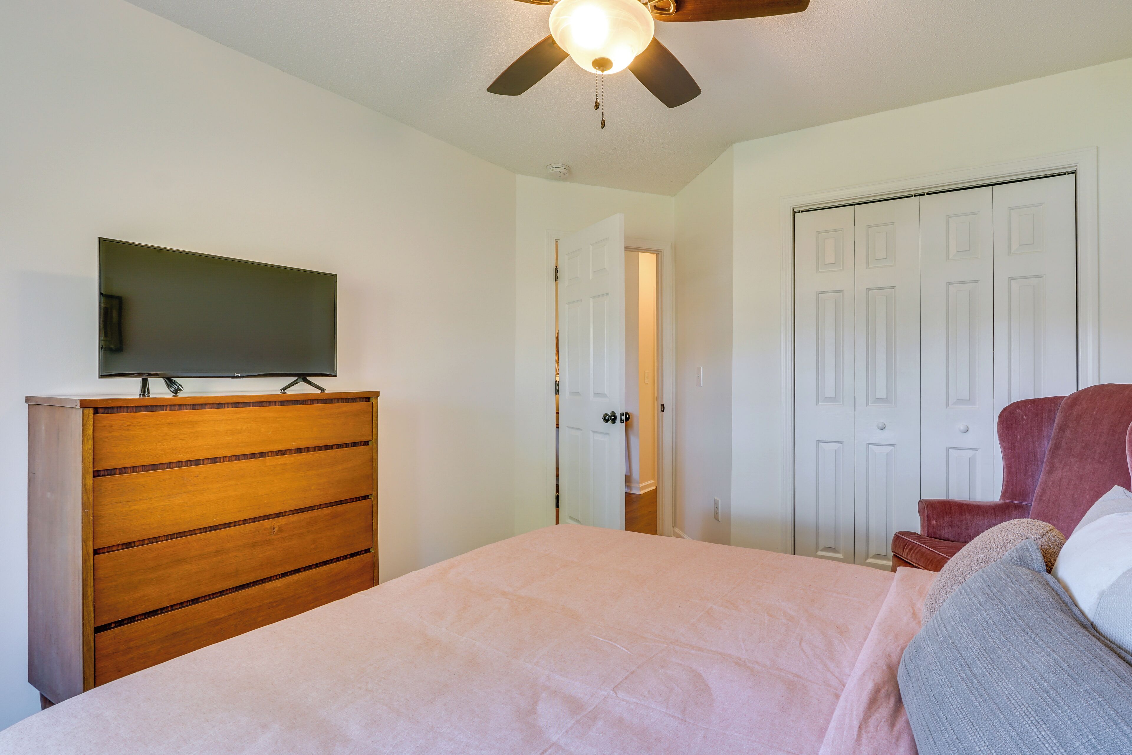 A bedroom with a bed, a wooden dresser, a television on the dresser, a ceiling fan, and a door.