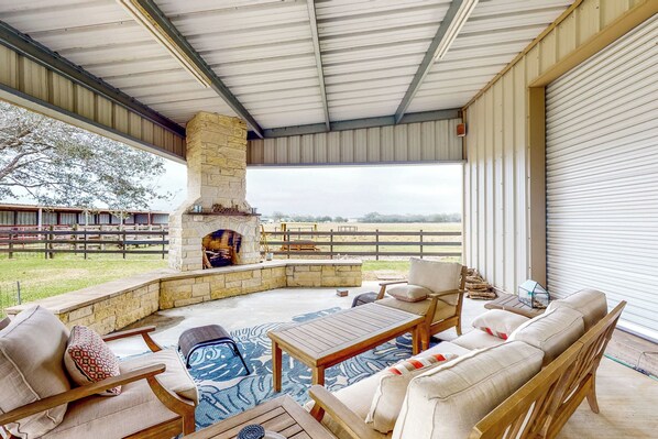 Un patio al aire libre con chimenea de piedra, mobiliario de madera y vistas a un campo cercado.