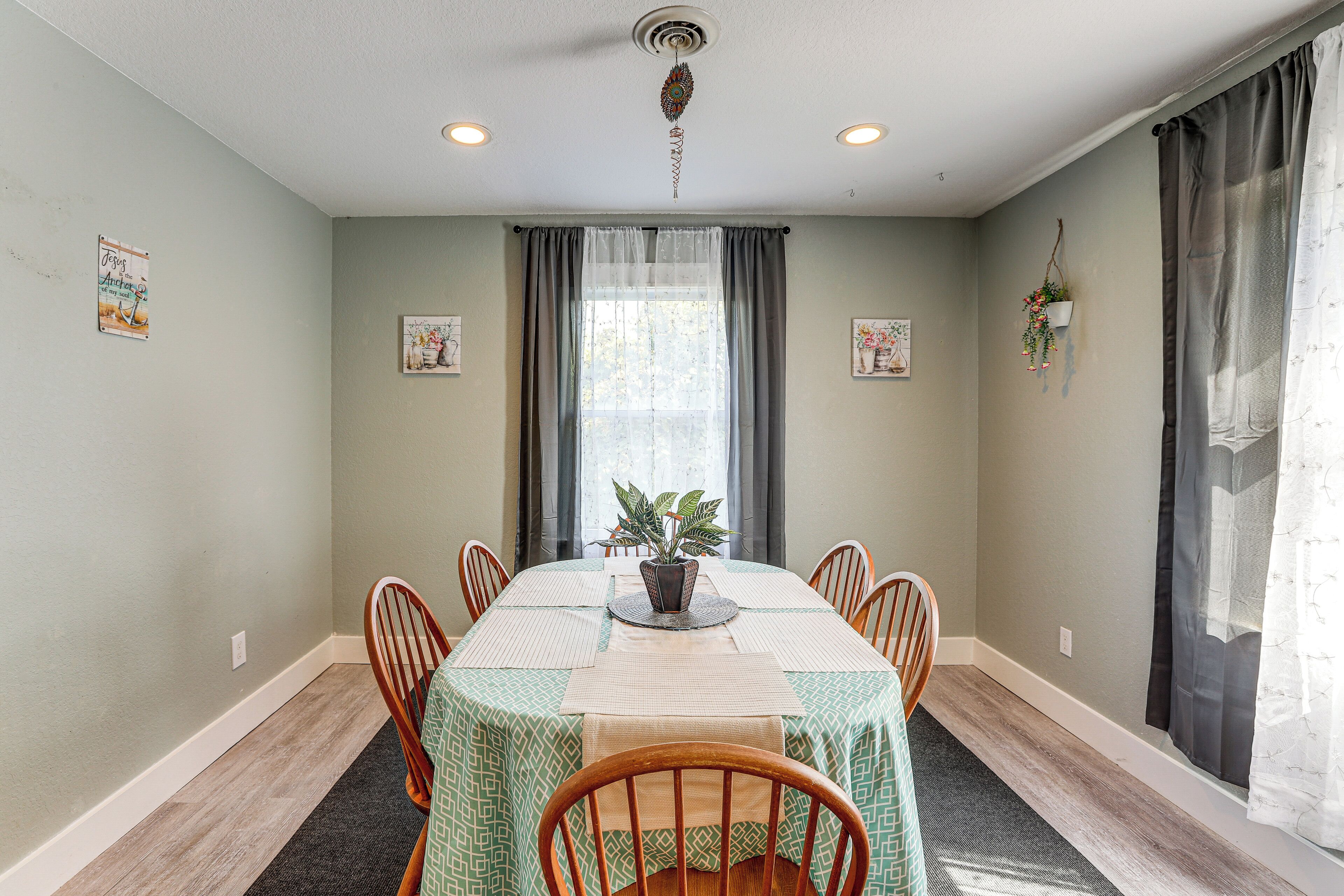 A dining room with a table set for six, a potted plant, and a window with curtains.