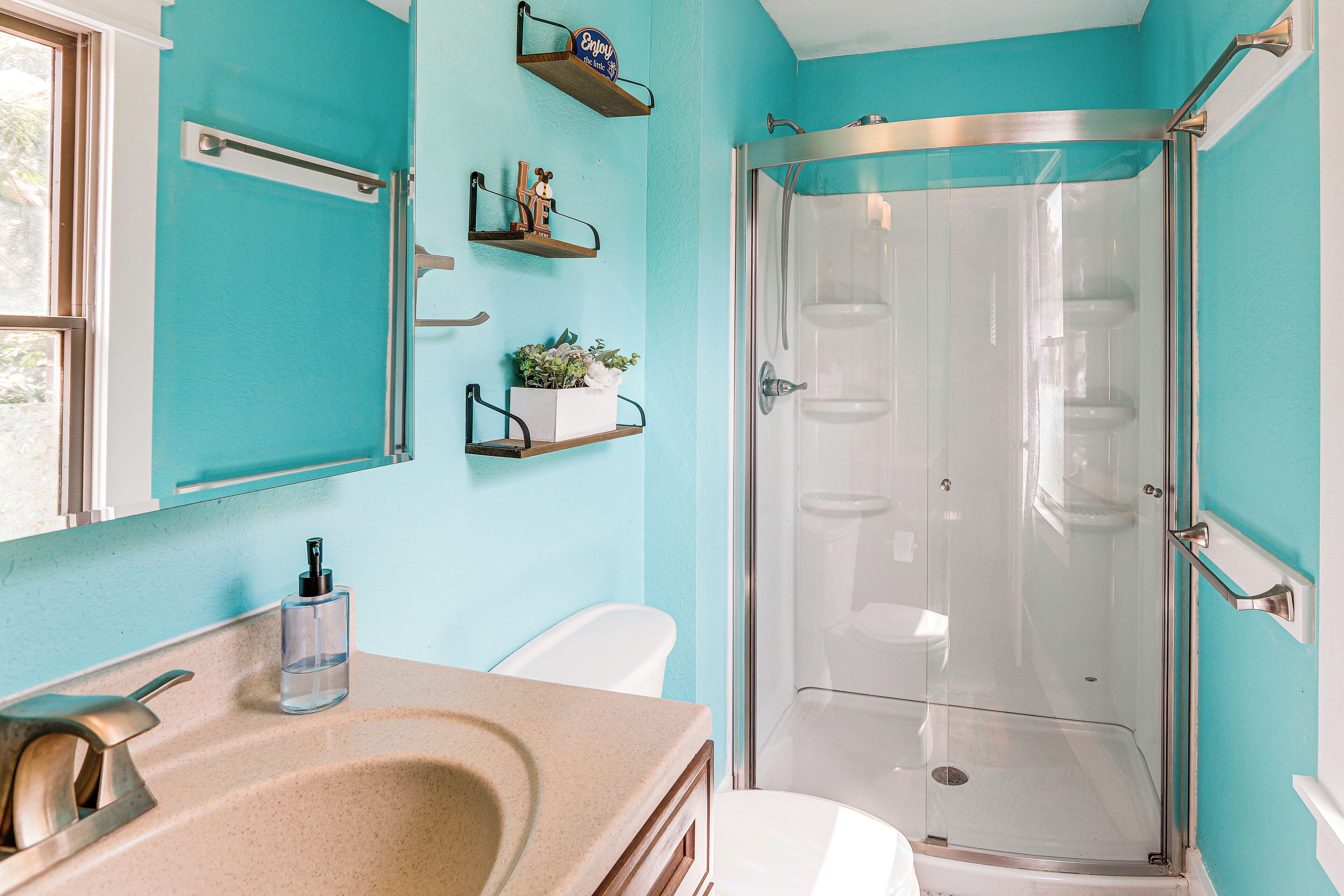 Une salle de bain avec une douche à parois de verre, un lavabo beige et des toilettes. Des étagères murales sont fixées au mur et accueillent des objets décoratifs.