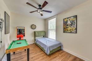 Una habitación con mesa de billar, ventilador de techo, dardos, máquina arcade Pac-Man y una cama.