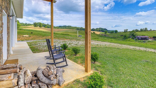 A covered porch with a bench, a stack of firewood, and a view of a green field and distant buildings. - Pet-friendly Home w/ Stunning Views in Sparta! (Sparta)