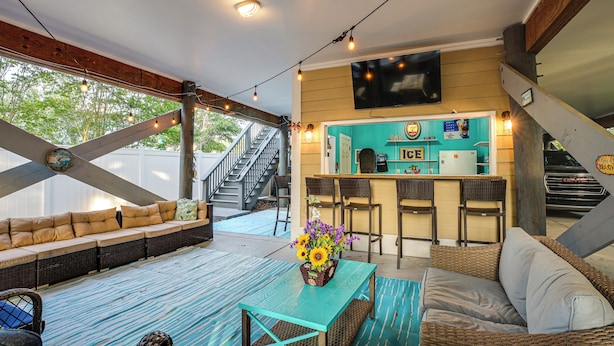 A covered outdoor seating area with a sofa, a table with a vase of flowers, and a bar area with chairs and a TV. - Shared Pool & Hot Tub! Seaside Murrells Inlet Home (Murrells Inlet)