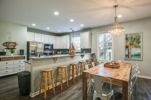 Una cocina con armarios blancos, una isla de madera con taburetes, una araña y un cuadro de una palmera.