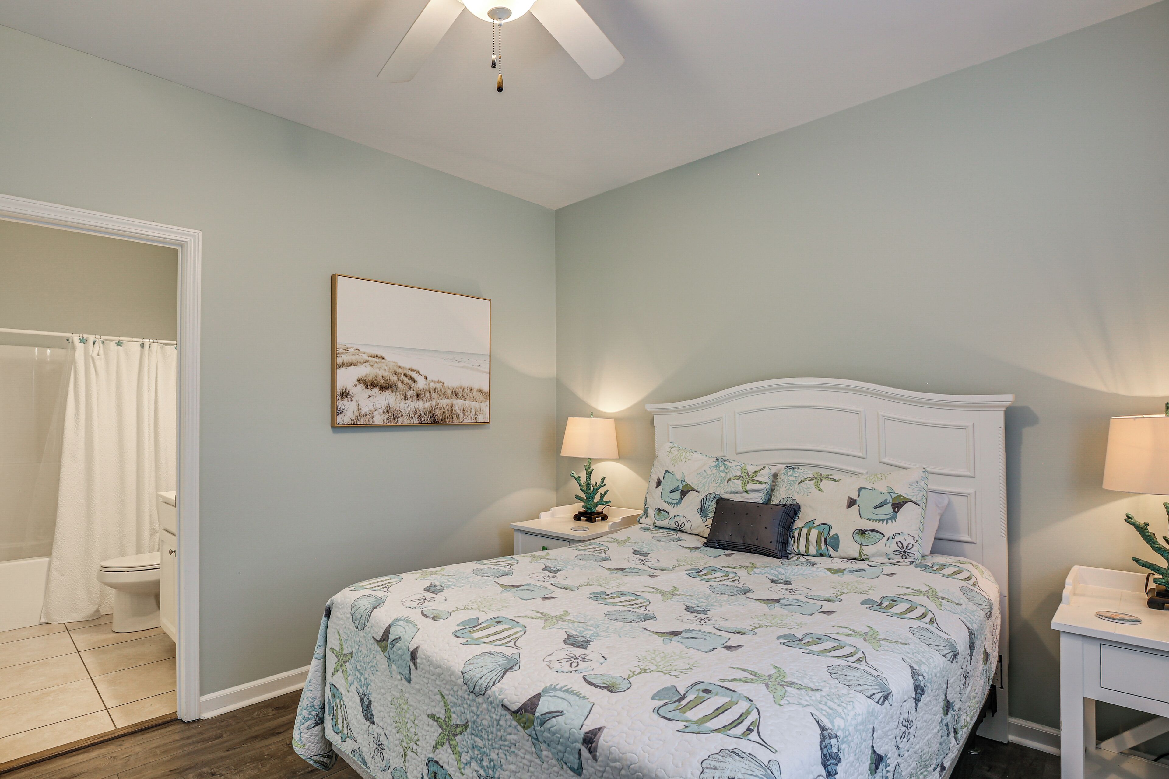 A bedroom with a bed, nightstands, a ceiling fan, a painting, and a bathroom with a shower curtain.