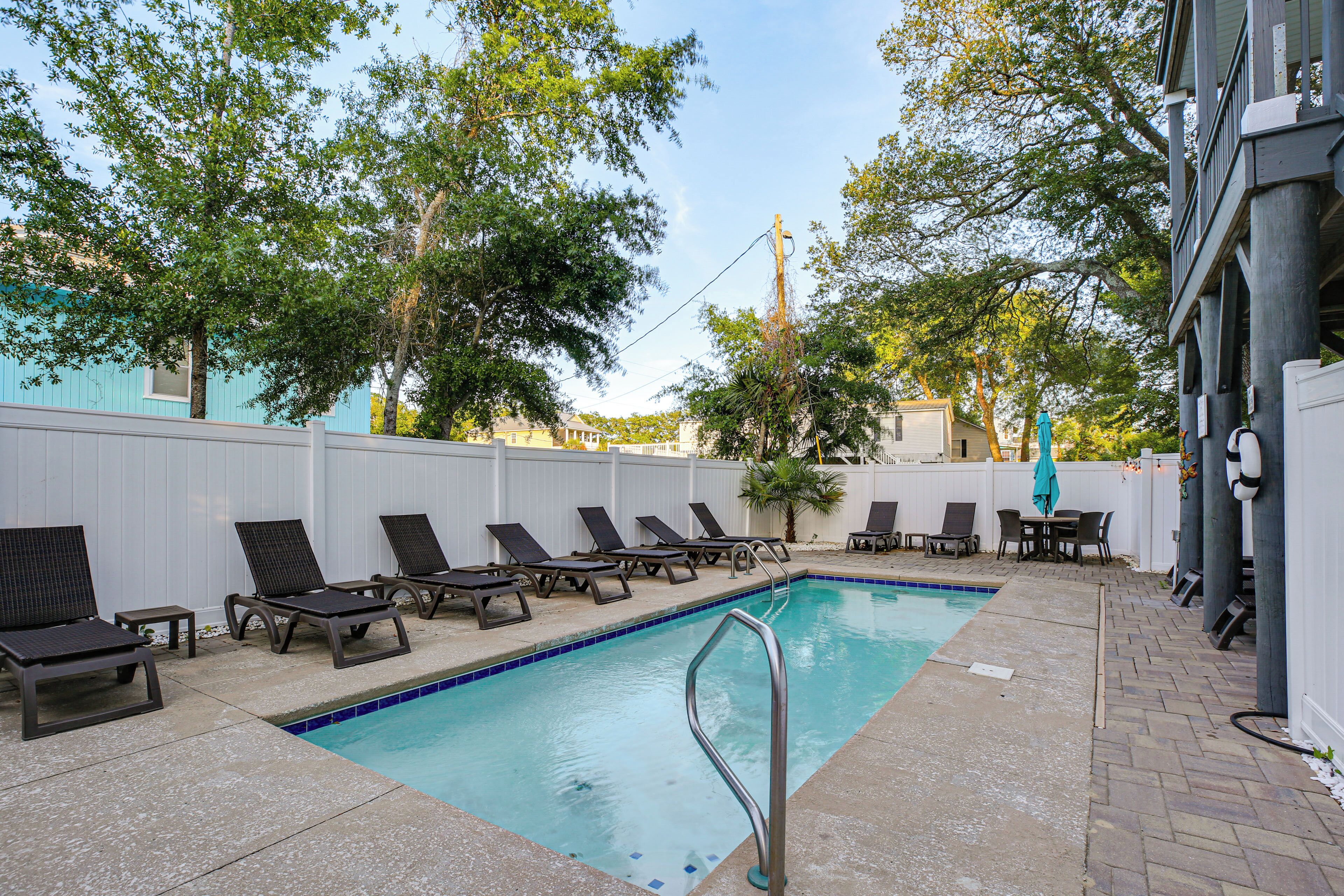 A swimming pool with lounge chairs and a table, surrounded by a white fence and trees.
