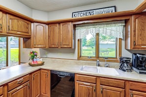 A kitchen with wooden cabinets, a dishwasher, a coffee maker, and a window with a view of a lawn.