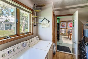 A laundry area with a washer and dryer, a window with blinds, a hanging rack, and a door leading to a room with a table and chairs.