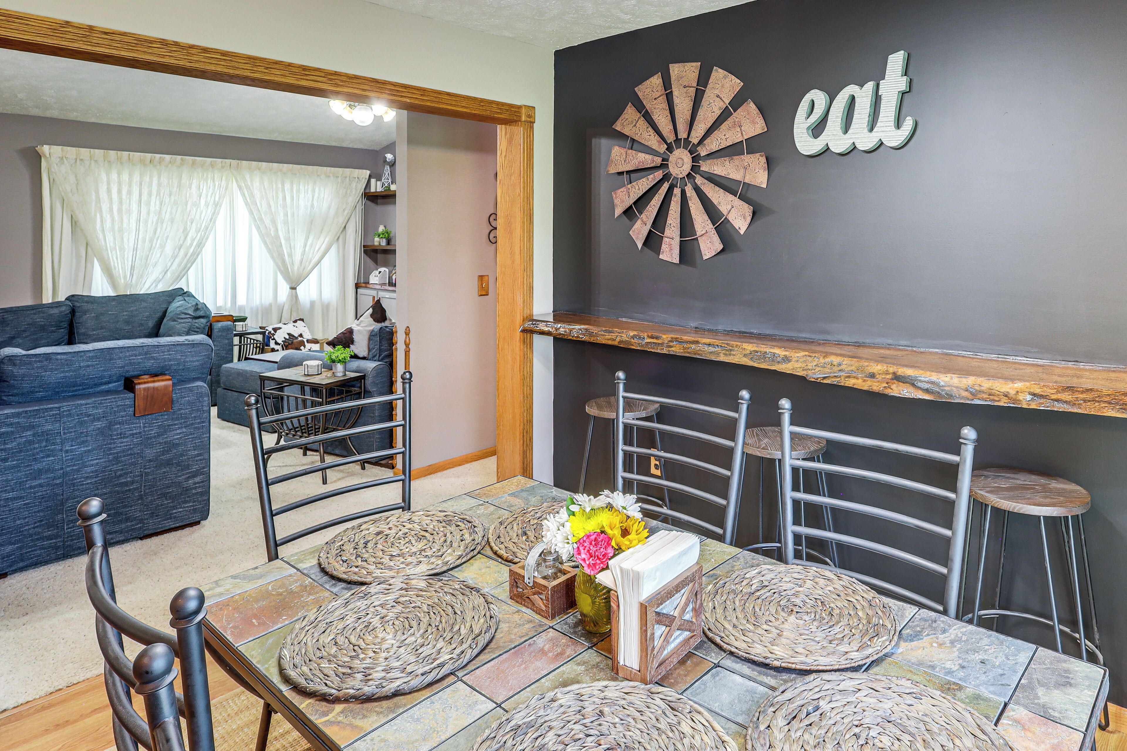 Un comedor con una mesa dispuesta para cuatro, rodeada de sillas metálicas, un adorno en la pared y un mantel individual tejido.