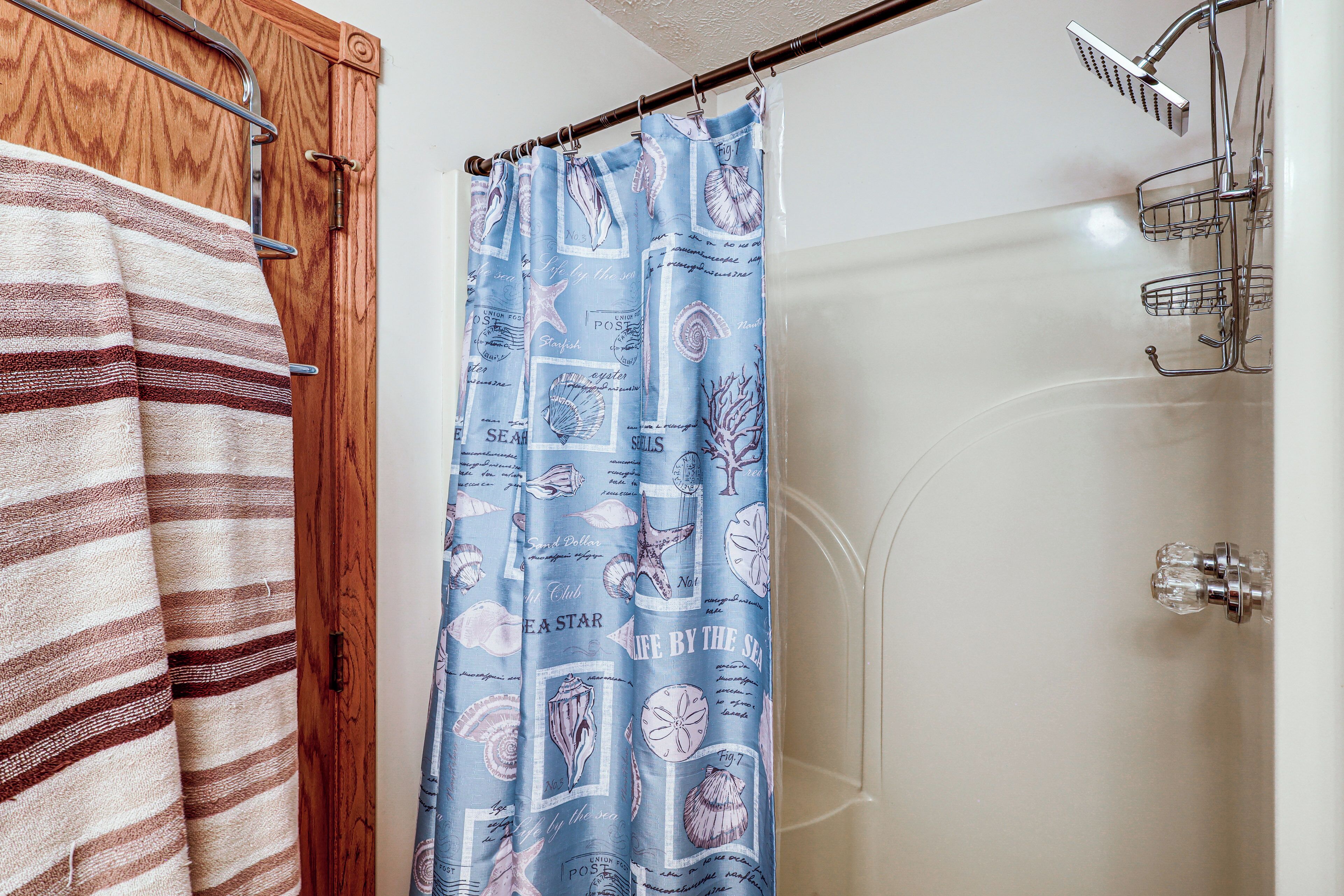 A bathroom with a shower curtain featuring sea shells and the phrase 'LIVE BY THE SEA,' a striped towel, and a wooden door.