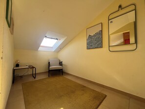 Une pièce dotée d’une lucarne, d’une chaise, d’une petite table avec une lampe, et de deux œuvres d’art encadrées accrochées au mur.