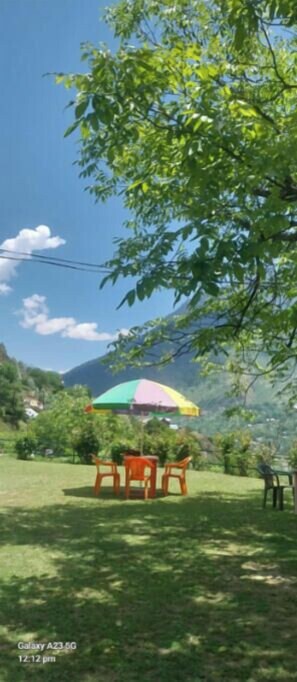 A grassy outdoor area with a table and chairs under a colorful umbrella, surrounded by trees and a mountain in the background. - Ahmed Lodge (Gund)