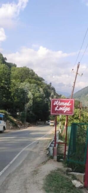 A sign for 'Ahmed Lodge' on a roadside with a view of a road and greenery in the background. - Ahmed Lodge (Gund)