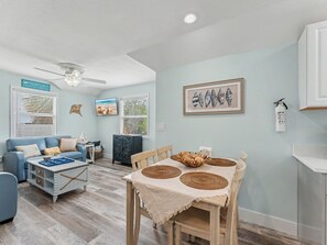 Ein Wohnzimmer mit einem blauen Sofa, einem Esstisch mit gewobenen Tischsets, einem Deckenventilator und einem gerahmten Kunstwerk mit Meeresschnecken an der Wand.