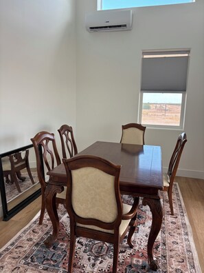 Uma sala de jantar com mesa e cadeiras de madeira, um tapete estampado, um aparelho de ar condicionado e uma janela com vista.