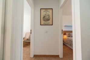 A hallway with a framed poster on the wall, a bed with a patterned bedspread in the background, and a chair with a cover. - Cozy Beach House (El Médano)