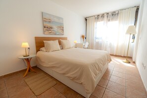 A hotel room with a bed, two bedside tables, a lamp, a painting on the wall, and a window with curtains. - Cozy Beach House (El Médano)