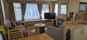 Interior compacto de una caravana con zona de estar, televisor, una mesita y kitchenette.