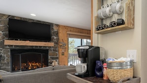 Ein Wohnzimmer mit Kamin aus Stein, einem Flachbildfernseher, einer Kaffeemaschine und einem Korbwägelchen auf einer Holzauslage.