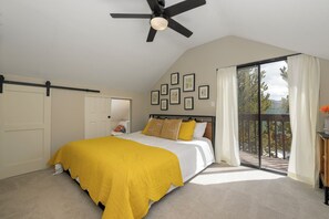 Quarto com cama, ventilador de teto, varanda com vista e quadros na parede.