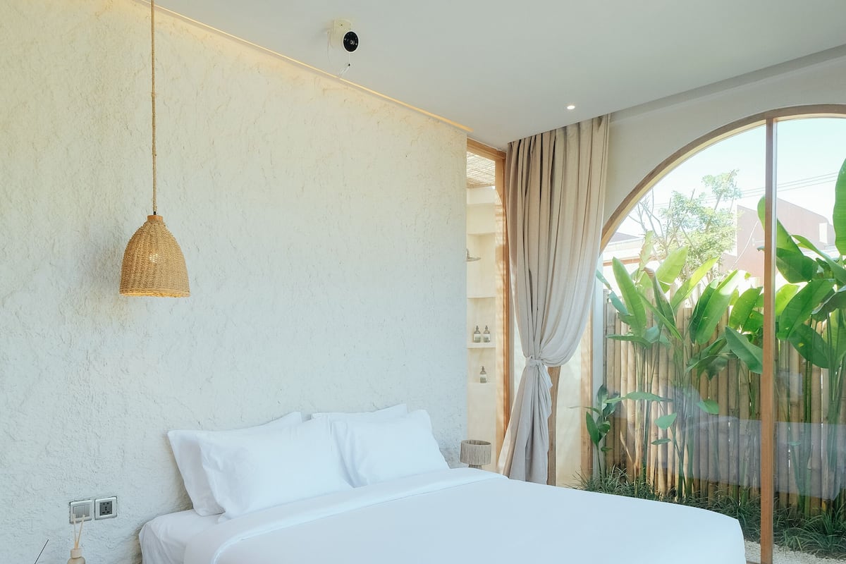 A modern bedroom with a wooden bed, a bedside table, a hanging light, and a view of greenery outside.