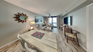 A dining area with a wooden table and chairs, a sunburst mirror, a painting, and a view through a window. - Harbour Place #108 | Lux Gulfside Condo, Partial Gulf View + Amenities (Orange Beach)