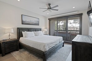 A bedroom with a large bed, a ceiling fan, a nightstand with a lamp, a desk, and a view of a snowy landscape through a sliding glass door. - Beaver Creek Slope-Side Luxury | UB306 (Beaver Creek)