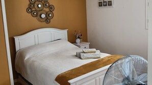 Ein Schlafzimmer mit Bett, Nachttisch, Wanddekoration und einem Standventilator.