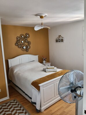 Un dormitorio con cama, mesita de noche, decoración en la pared y un ventilador de pie.