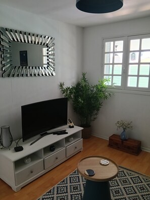 Ein modernes Wohnzimmer mit einem Flachbildfernseher, einem Ziertepiegel, einer Topfpflanze, einem kleinen Tisch und einem Fenster mit Jalousien.