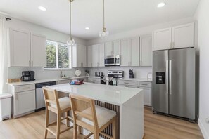 A modern kitchen with white cabinets, stainless steel appliances, a central island with bar stools, and pendant lighting. - Theater Room, Game Room & Family Fun near Disney (Davenport)