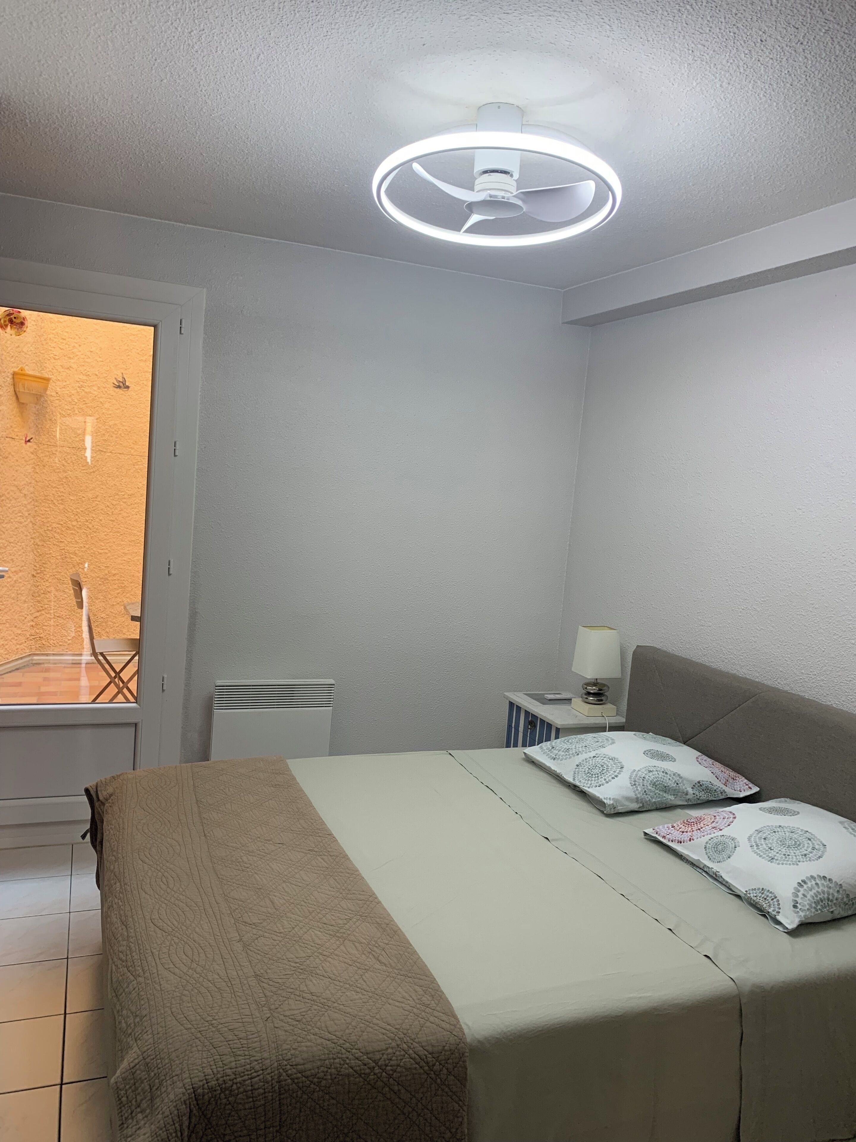 A bedroom with a bed, a bedside table with a lamp, a ceiling fan, and a window with a view of the outside.