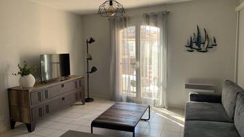 Un salon avec un téléviseur, une plante, un lampadaire, un canapé et une fenêtre avec des rideaux.
