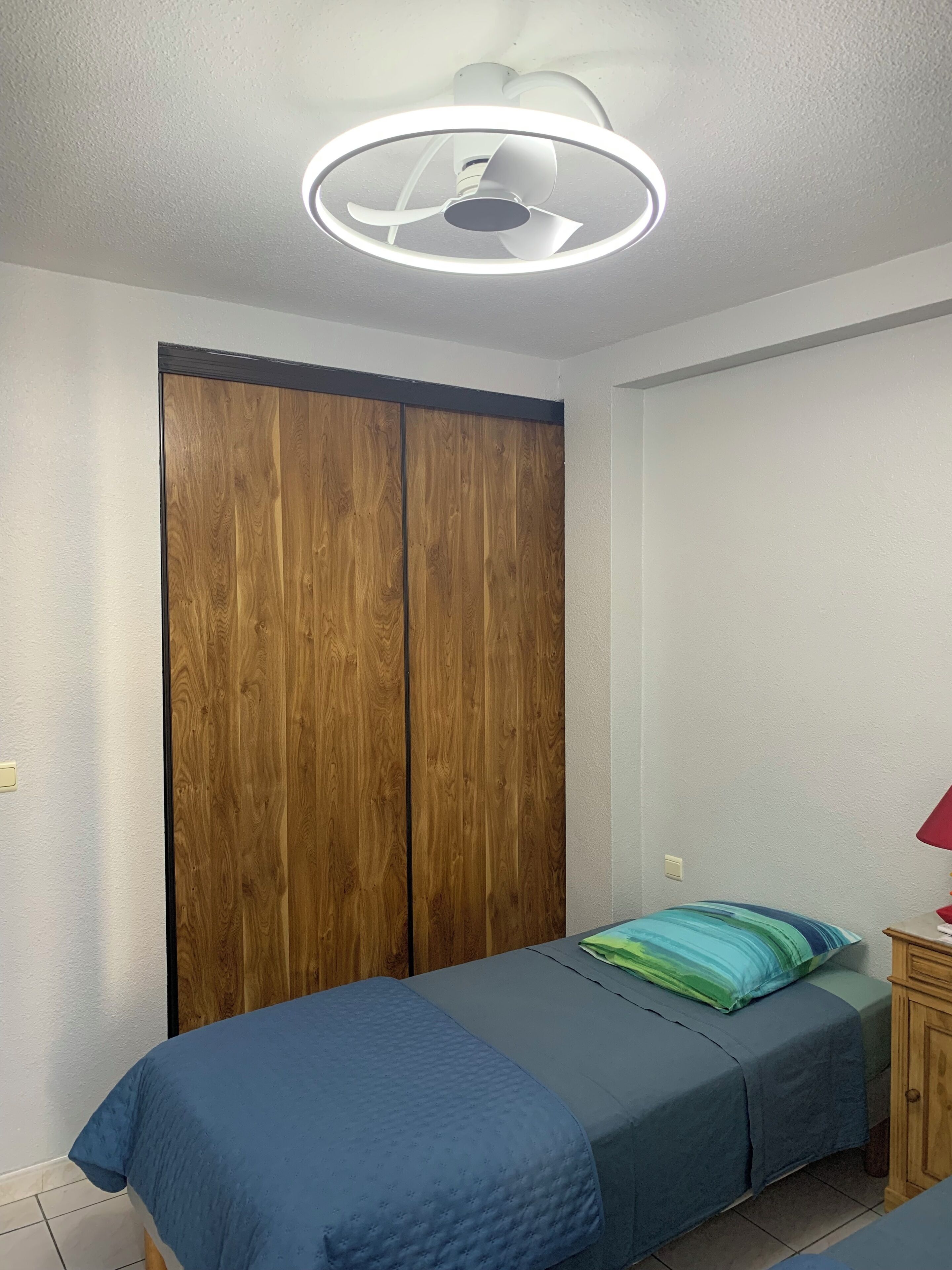 Ein Schlafzimmer mit einem Bett, einem Kleiderschrank, einem Nachttisch mit Lampe und einem Deckenventilator.
