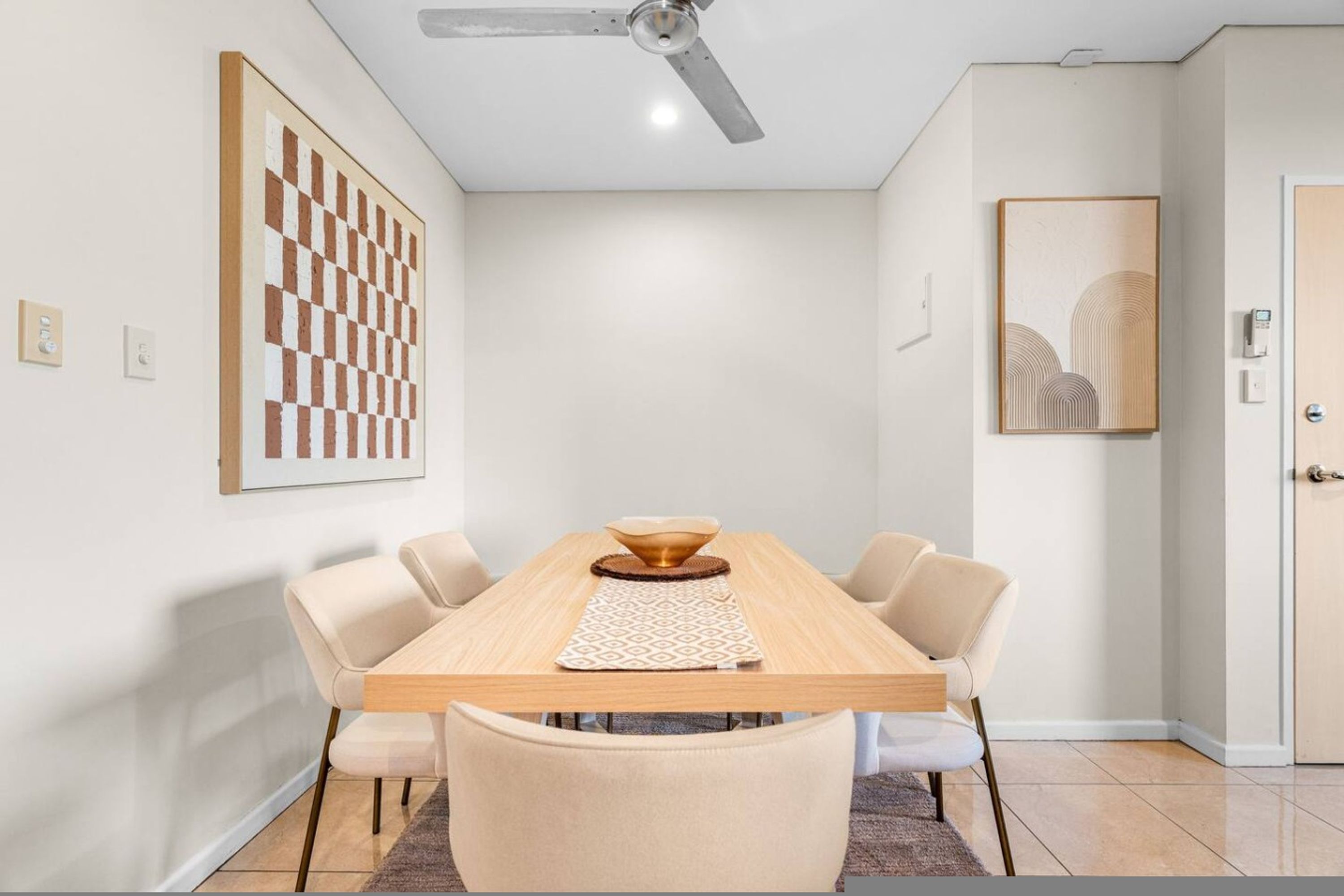 A dining area with a wooden table, beige chairs, a ceiling fan, and artwork on the walls.