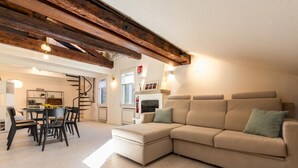 Ein modernes Wohnzimmer mit einer großen, beigefarbenen Eckcouch, Holzbalken an der Decke, einem Essbereich mit Tisch und Stühlen sowie einer Küche im Hintergrund.