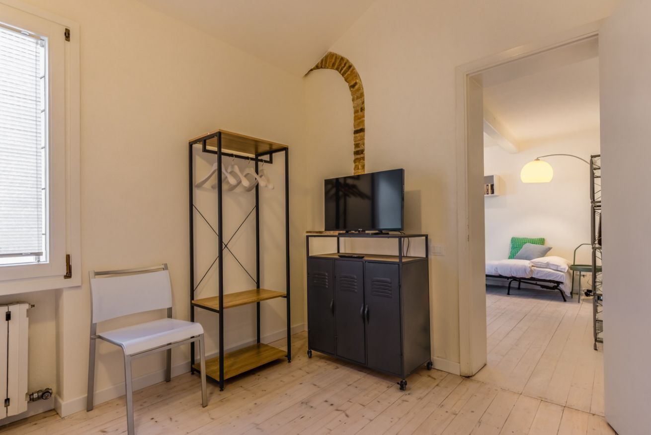 A room with a white radiator, a white chair, a metal shelving unit, a TV on a stand, and a bed with a green pillow.