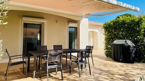 Une terrasse extérieure avec une table et des chaises, un barbecue abrité et un bâtiment à la façade beige.