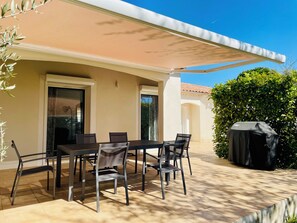 An outdoor patio with a table and chairs, a covered grill, and a building with a beige facade. - Large Provencal house (Pujaut)