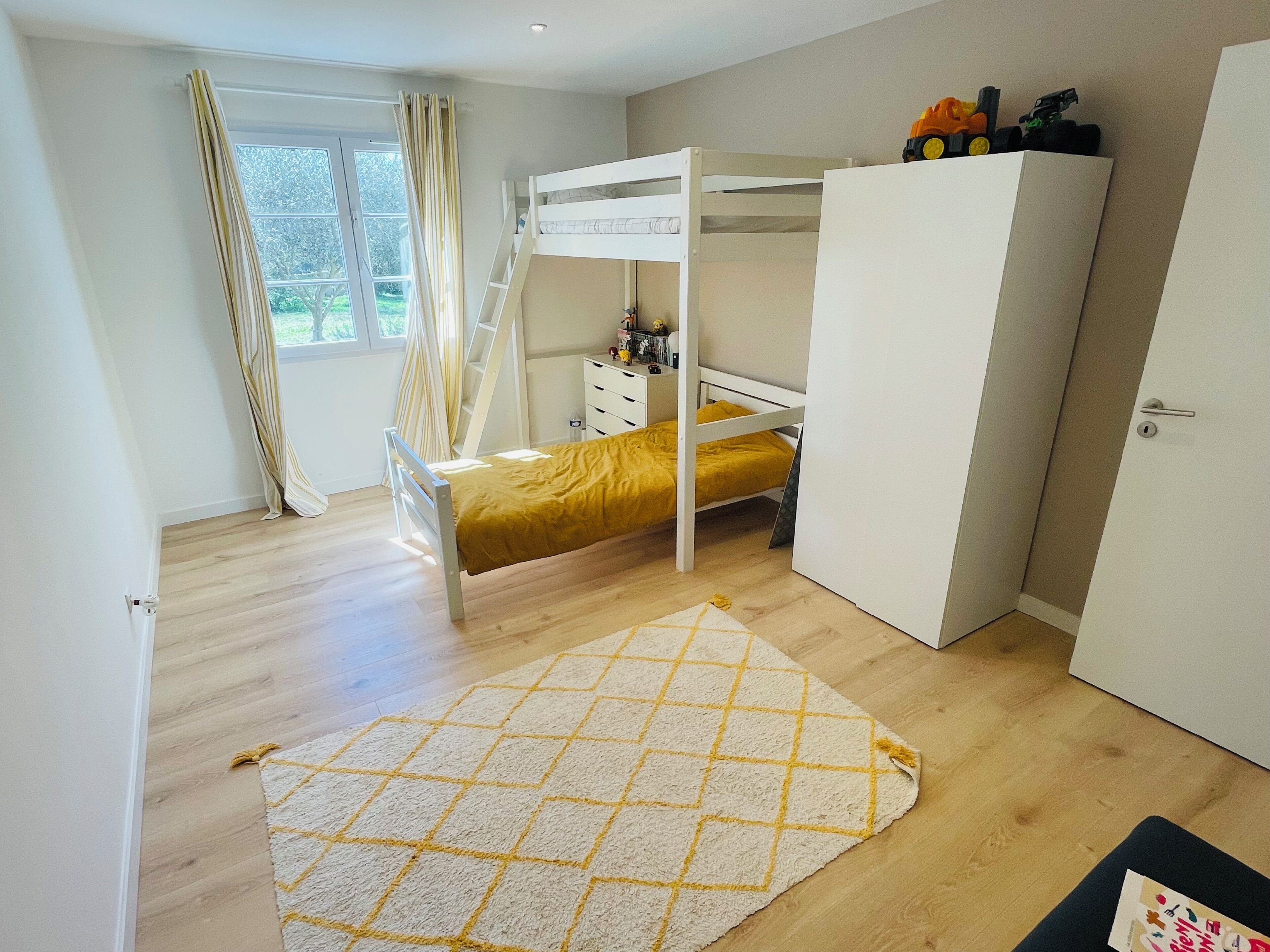 A bedroom with a bunk bed, a wardrobe, a desk, and a window with curtains.
