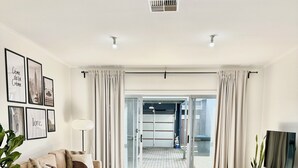 Een moderne woonkamer met een ronde eettafel, witte stoelen, een bank en een televisie. Er hangen ingelijste foto's aan de muur en staat een potplant.