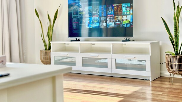 Een moderne woonkamer met een flatscreen televisie op een witte kast, houten vloeren en twee potplanten.