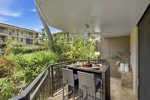 A balcony with a table set for dining, surrounded by lush greenery and a view of other buildings. - Kona Makai 3-105: Ground Floor, Ocean View Beauty (KAILUA KONA)